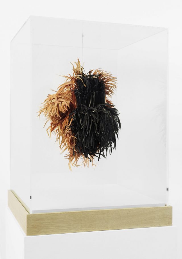 An abstract sculpture made of sliced pages of books hangs in a glass vitrine. The half black and half straw-colored slices look like a mass of hay