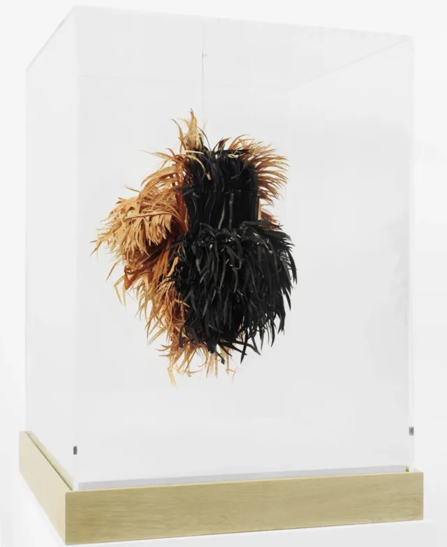 An abstract sculpture made of sliced pages of books hangs in a glass vitrine. The half black and half straw-colored slices look like a mass of hay