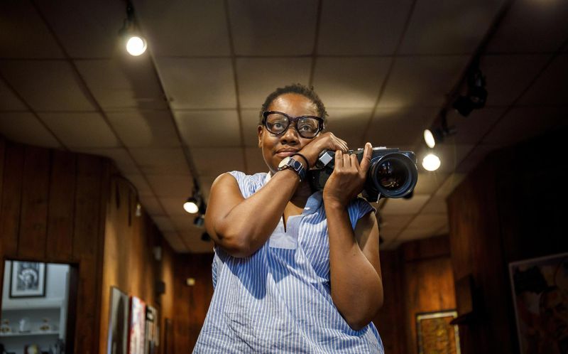 An African American woman with short cropped hair and black-framed glasses holds a large camera to the right of her face. The room she is in is dimly lit and wood-paneled. 