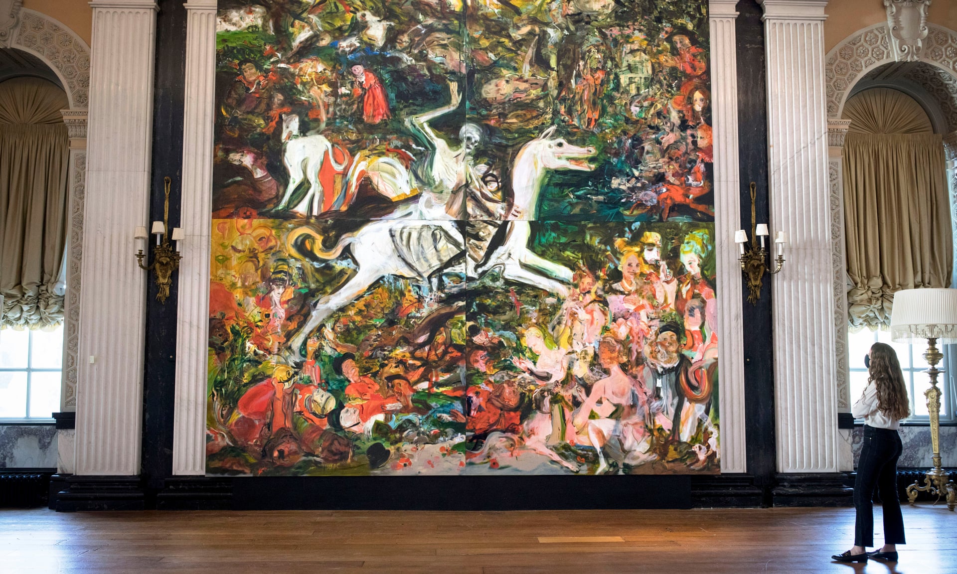 In a large palace room, a woman gazes up at a floor-to-ceiling mural. In it, a skeleton with its right hand raised, rides on a skinny, deformed white horse in the middle of four panels. Abstract light-skinned human figures sit around the horse’s feet, some lounging and speaking amongst themselves. Streaks of gold, green, and teal surround the figures.
