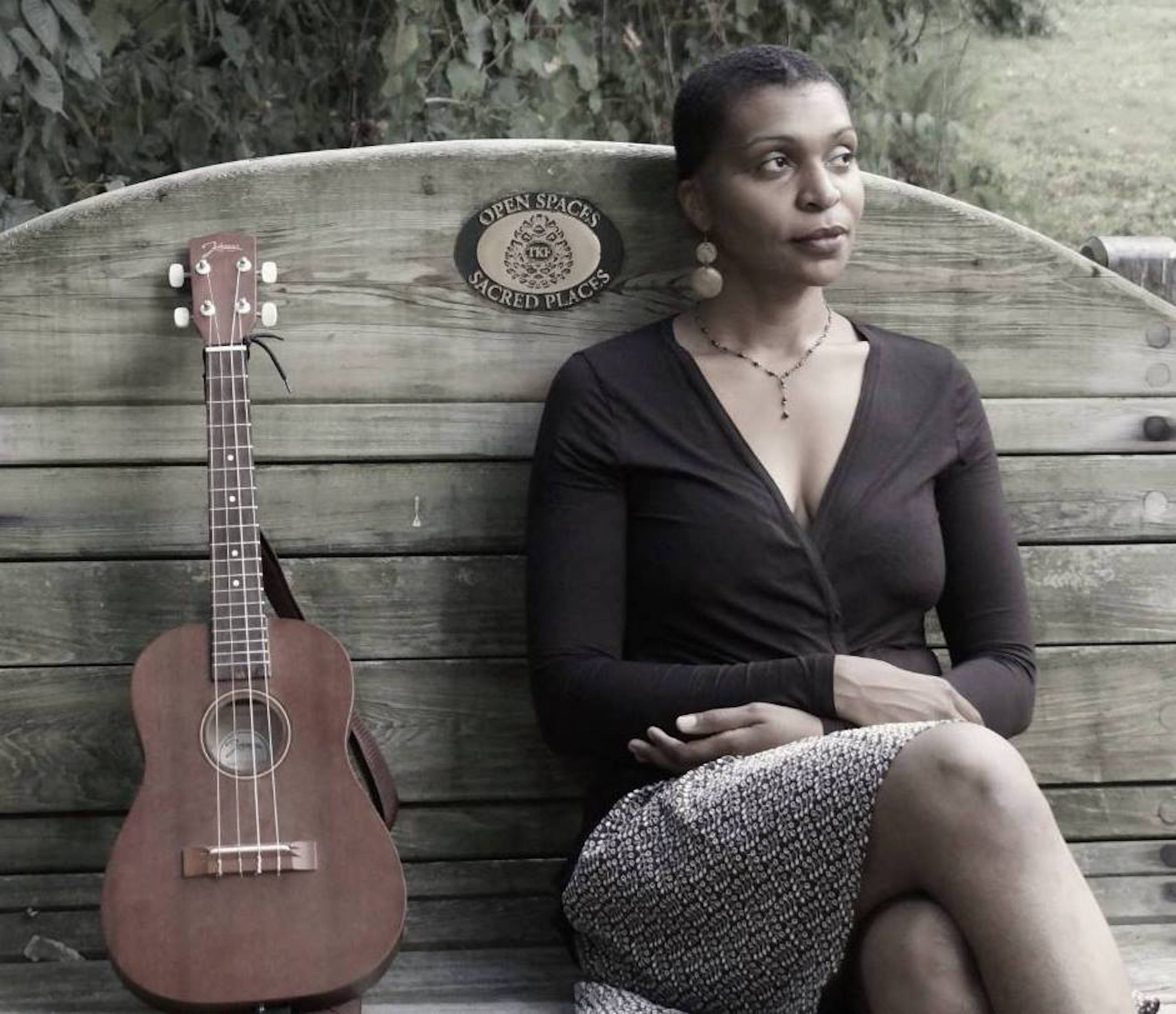 Courtney Dowe sits on a wooden bench with a high back with a standing brown guitar on her right. Her legs are crossed and her arms are crossed on her lap. She wears a black top, a patterned skirt, earrings, and a necklace, and she has very short hair. Behind her is lush greenery.