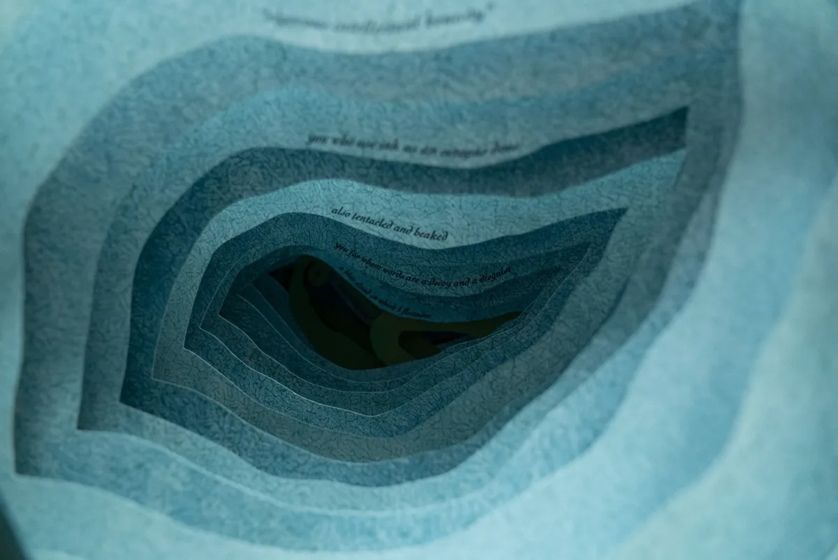 A close up photograph of an artist's book. Each layer of blue paper has an organic, irregular shape cut out of the middle so the layers form a tunnel. A line of text is printed in small type on each page, receding into the tunnel like an underwater cavern.