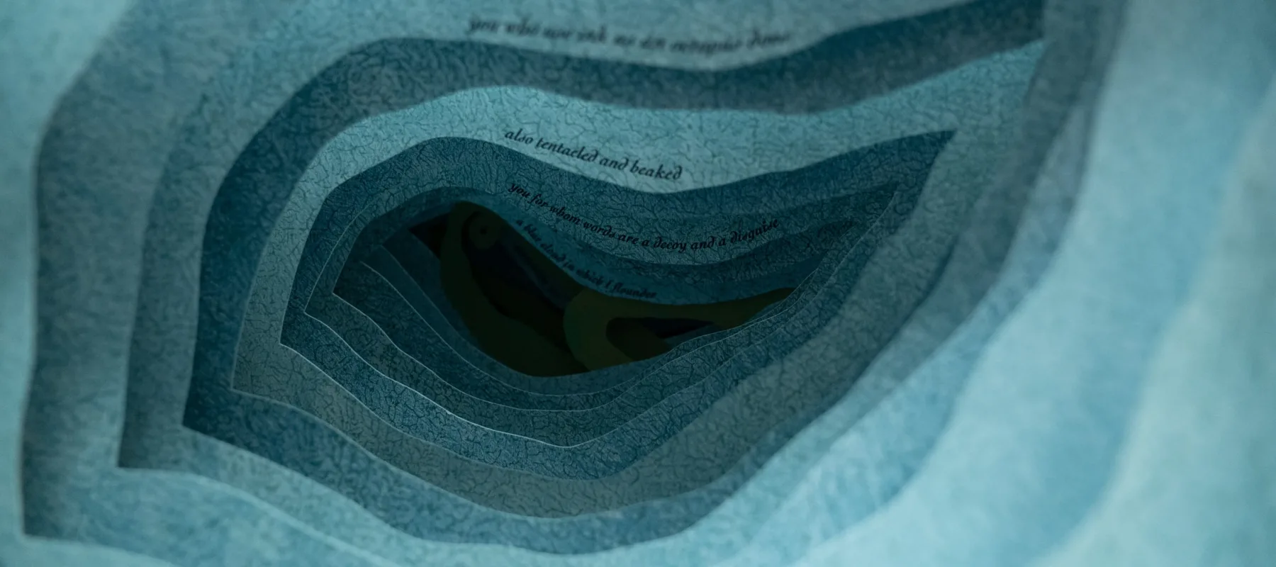 A close up photograph of an artist's book. Each layer of blue paper has an organic, irregular shape cut out of the middle so the layers form a tunnel. A line of text is printed in small type on each page, receding into the tunnel like an underwater cavern.