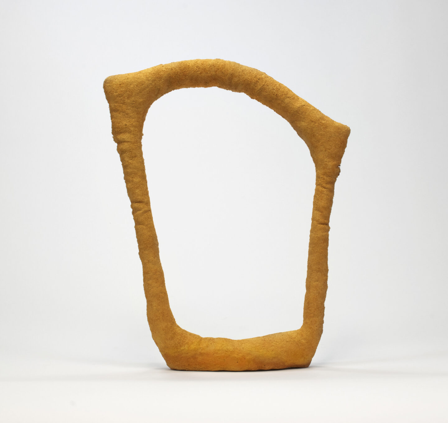 A trapezoidal abstract orange sculpture made of paper pulp with a top arch that connects the two sides. The sculpture is thin with a big void space in between the four sides.