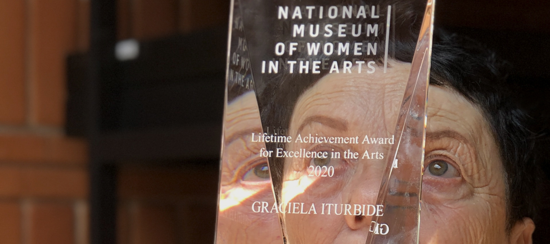 A light-skinned older woman with brown cropped hair holds a clear rectangular award up to her face, so that half of her face is visible through the award; sunlight streams in on the woman's face and hands.