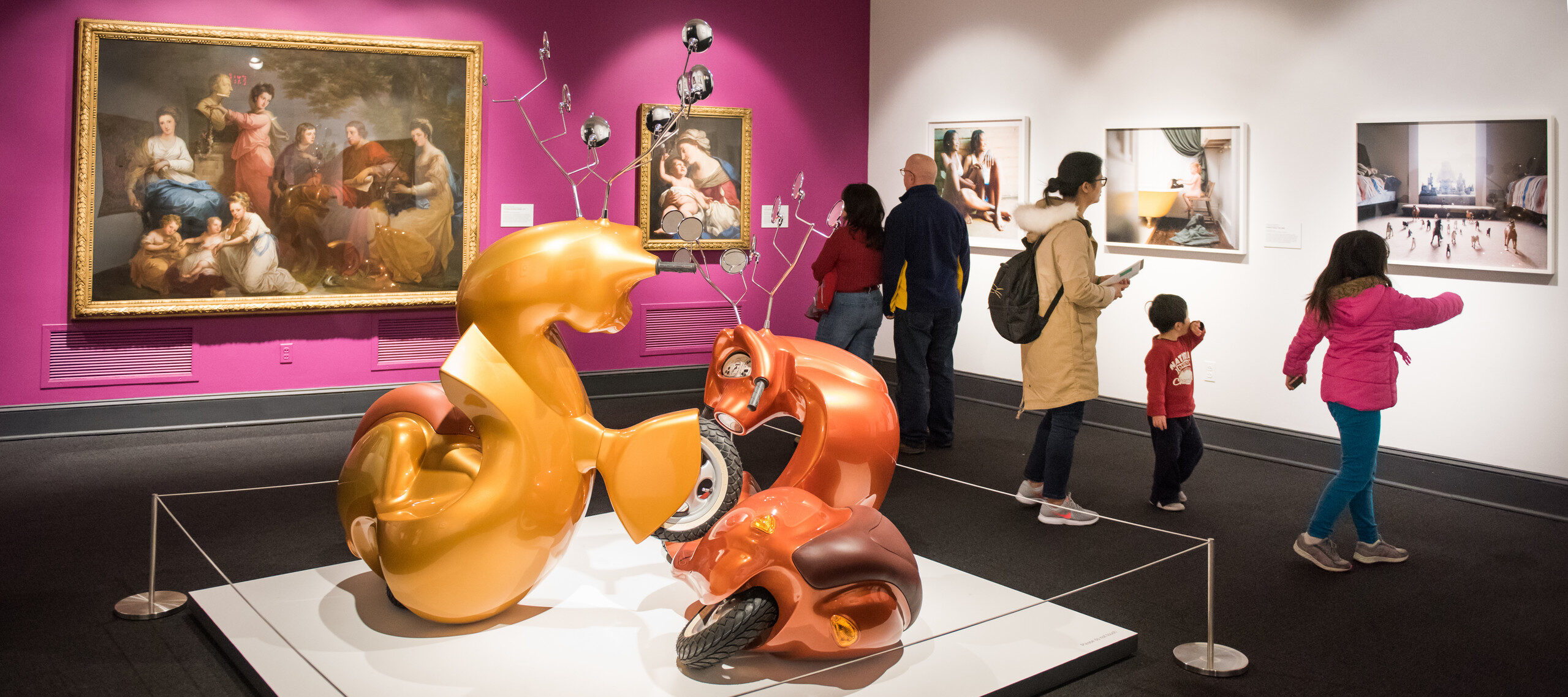 View of a gallery with walls painted magenta on the left and white on the right. In the middle is an installation of two stylized scooters that look like two deer interacting. To the right, three adults and two children look at different artworks mounted on the wall.