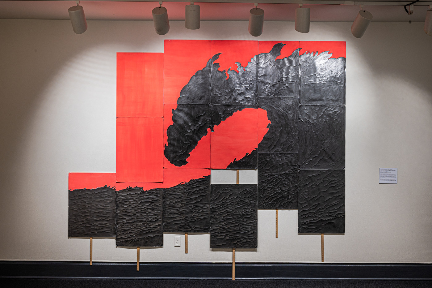 Six rectangular panels of varying heights with a coral background are hung together on a wall with yardsticks attached to the bottoms of each panel. Each panel includes a part of a textured graphite drawing that looks like a giant crashing wave when seen as a whole.