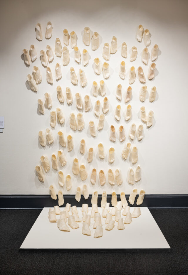 A contemporary art installation made up of countless beige foot sculptures that extend from the walls onto a low white platform on a dark floor. The feet are in pairs in varied sizes and staggered as if moving.