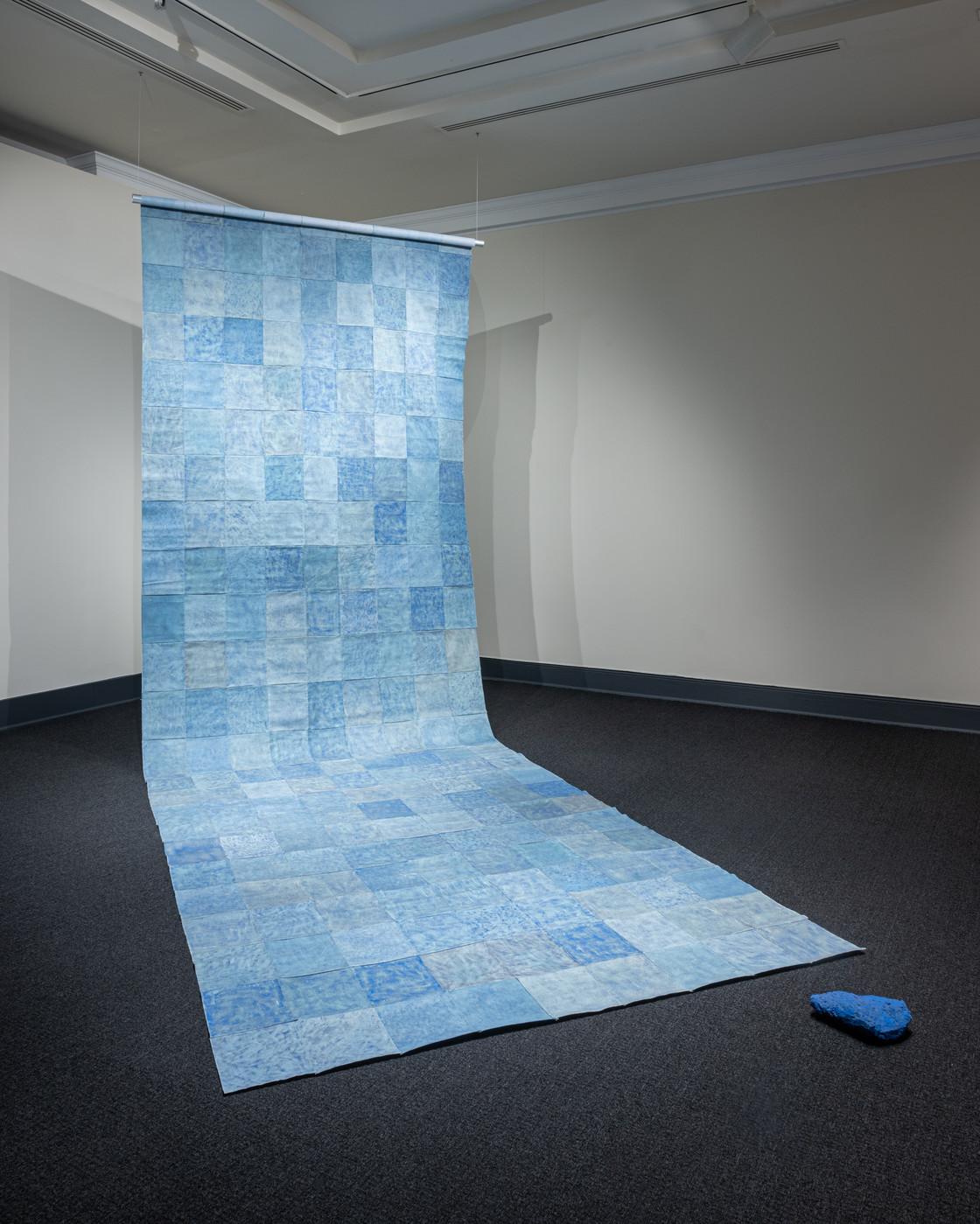 A floor to ceiling contemporary installation of blue square sheets patched together into a long panel that hangs down from two wires and partly rests flat on the floor. A dark blue object lays nearby. 