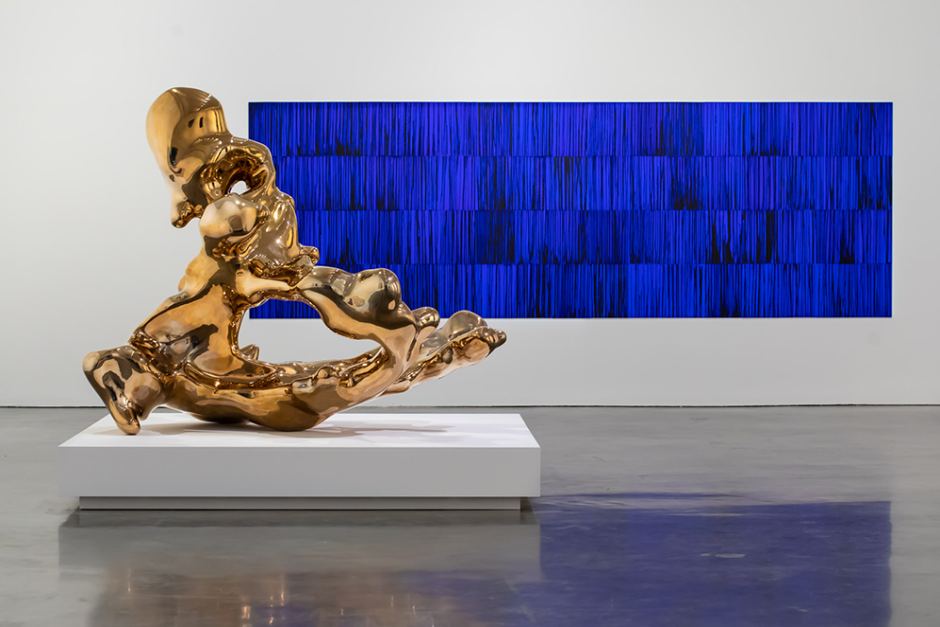 On the left, a large, gold metallic abstract sculpture reflects the light of the gallery. The sculpture is an uneven liquid bronze triangle with a hole in the middle. In the background sits a wide rectangular painting of blue and black upward brush strokes.