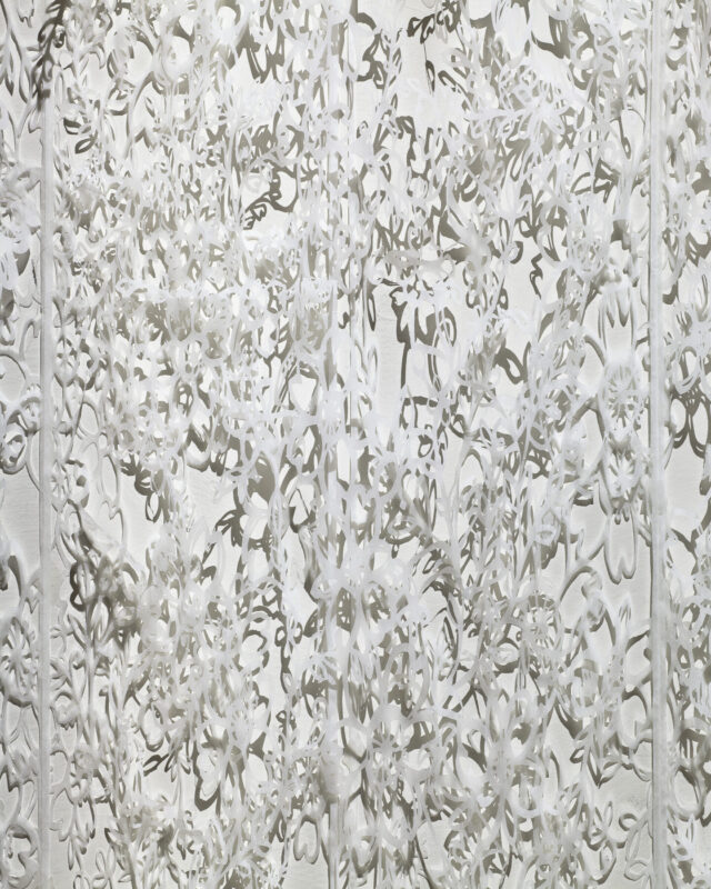 Detail view of delicate, white tissue paper that has been cut with intricate floral patterns which create shadows on the wall behind it.