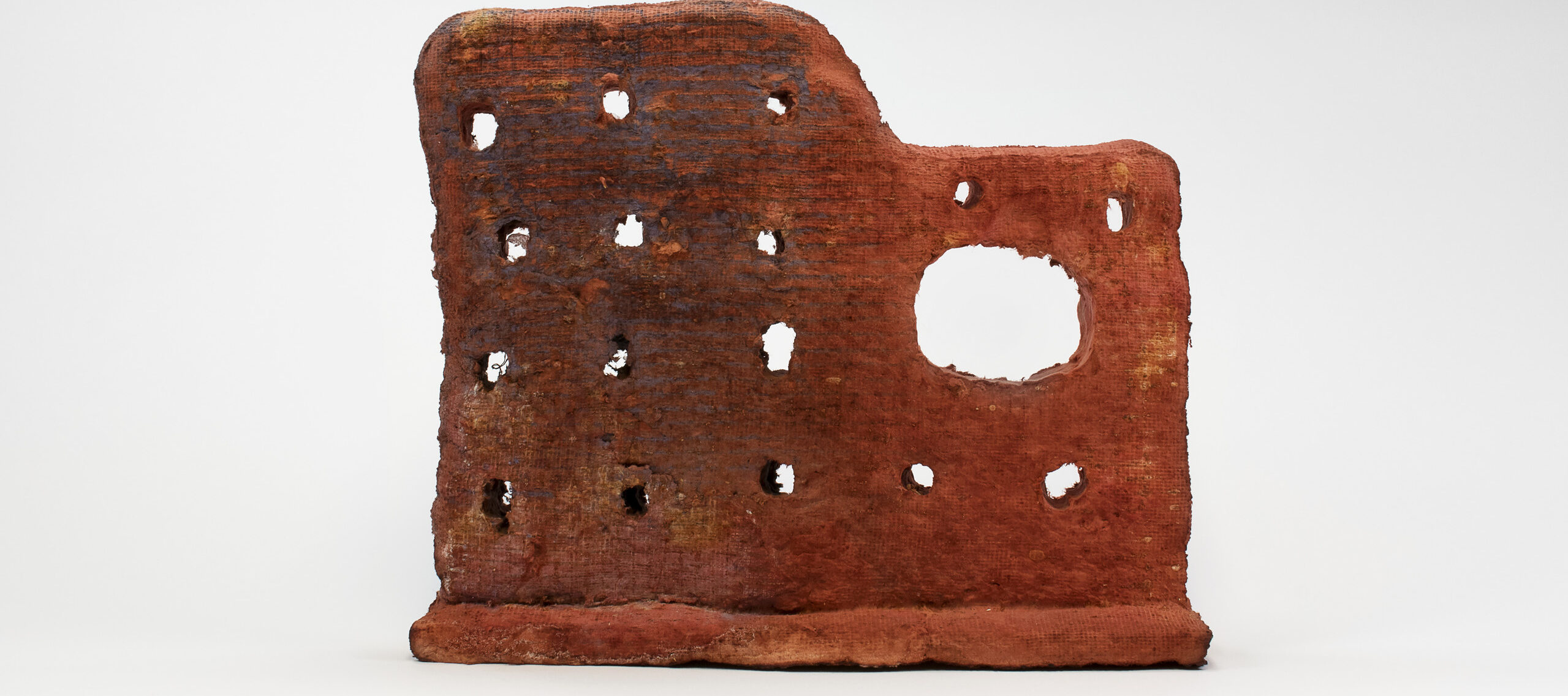 An irregular shaped red abstract sculpture that looks like the side wall of an old building structure. Rows of holes and one bigger void space cover the surface of the grainy, rough sculpture.