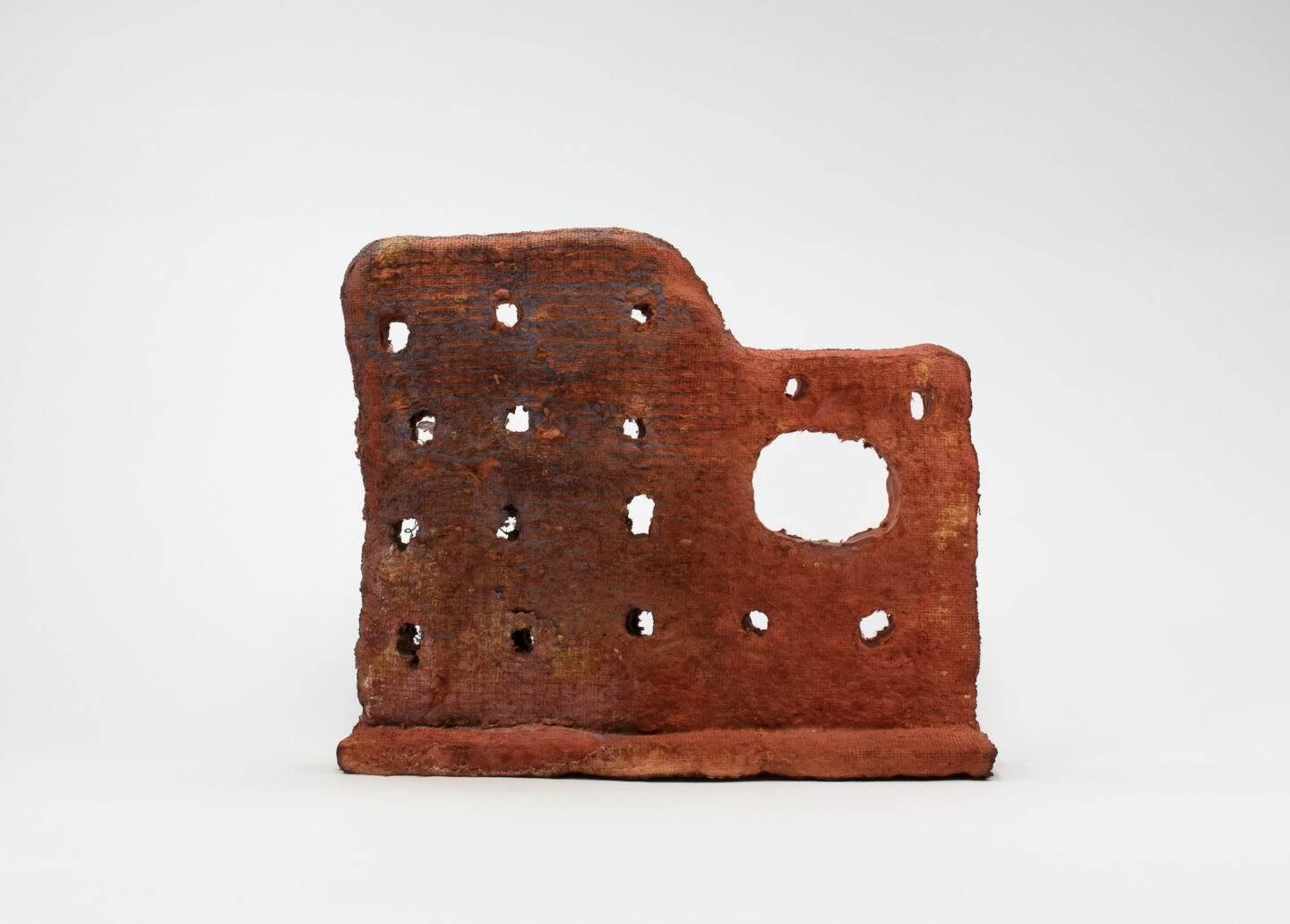 An irregular shaped red abstract sculpture that looks like the side wall of an old building structure. Rows of holes and one bigger void space cover the surface of the grainy, rough sculpture.