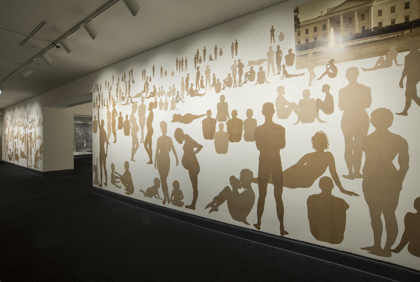 A long rectangular wall with many silhouetted figures made out of brown Kraft paper. The figures are of men, women and children in different positions as if in a landscape. They are different sizes to indicate foreground, middle ground and background. A sepia-toned photograph of the White House at the upper right corner.