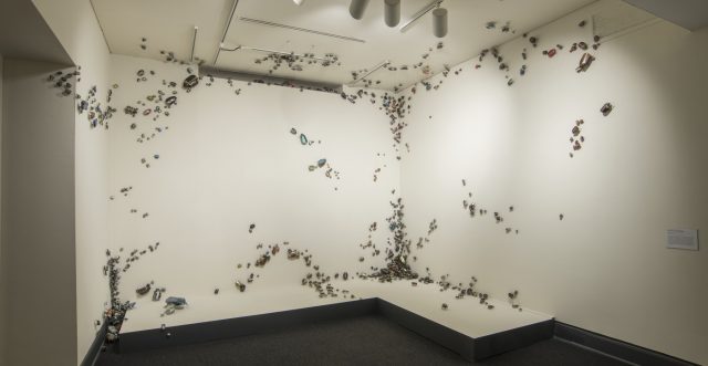 Installation view of thousands of small insects swarming out onto white walls, ceiling, and onto the dark floor.