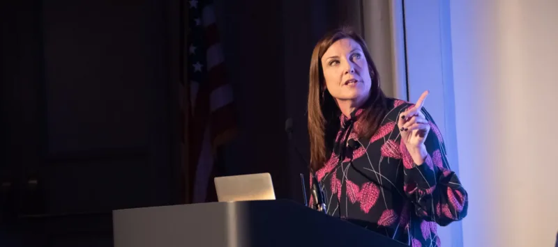 Jackie Payne—a light-skinned adult woman with straight, dark brown hair—stands at a podium in front of a laptop, looking to her side and behind her at a projected presentation she’s giving. Her left hand is raised to point at the presentation while she speaks.