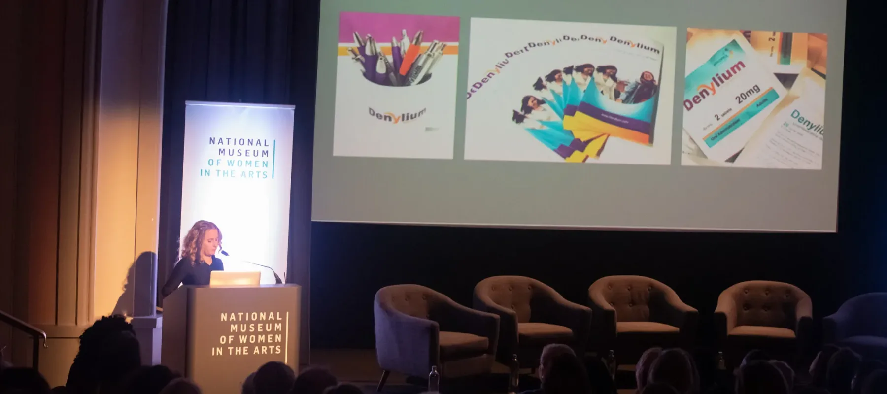 Kim Loper speaks at a podium on the stage of a NMWA Fresh Talk event. She is a light-skinned adult woman with curly, shoulder-length blonde hair, and is wearing a black top. Behind her is a projected presentation featuring flyers, brochures, and a mug full of pens that all say "Denylium."