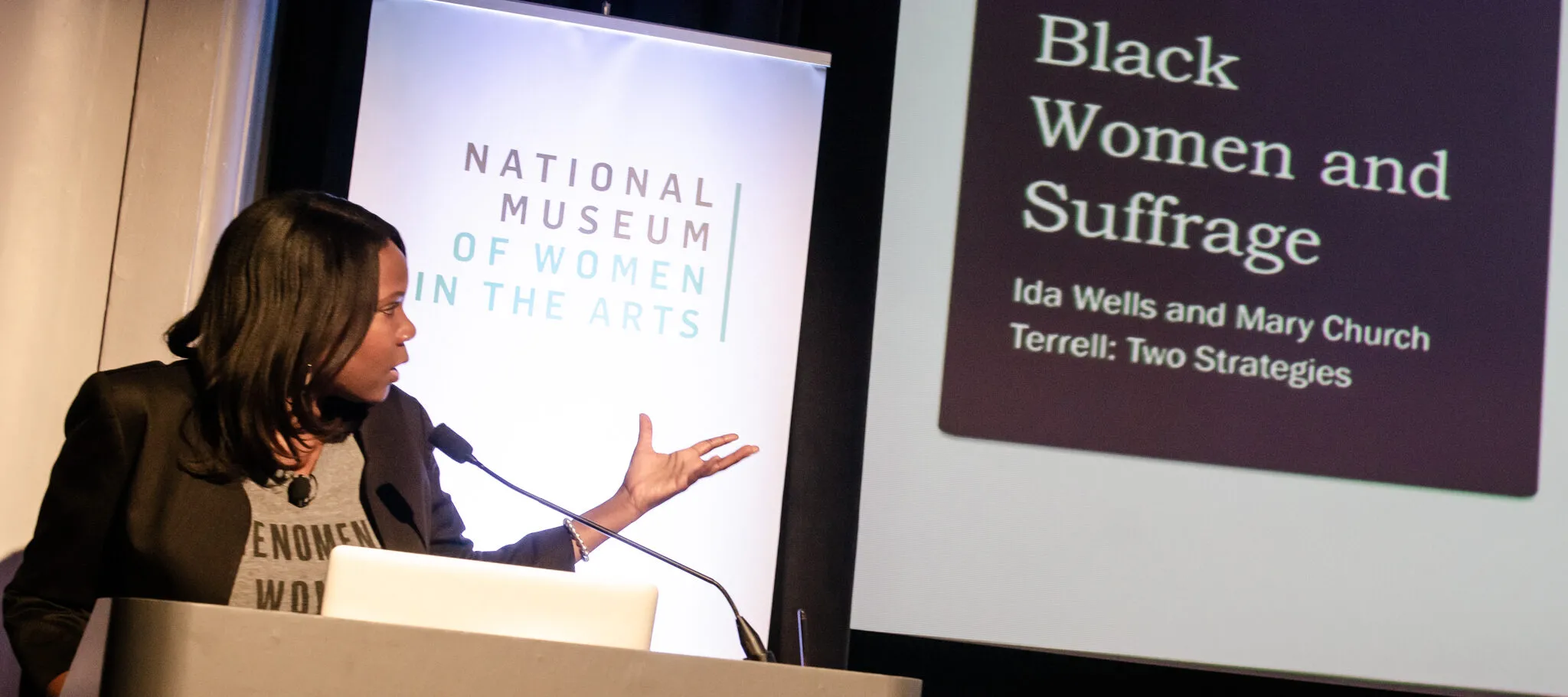 Adjoa B. Asamoah—a dark-skinned adult woman—stands at a podium with the NMWA logo on the front. She speaks into a microphone while gesturing at a projected presentation slide behind her that reads “Black Women and Suffrage. Ida Wells and Mary Cherch Terrell: Two Strategies.”