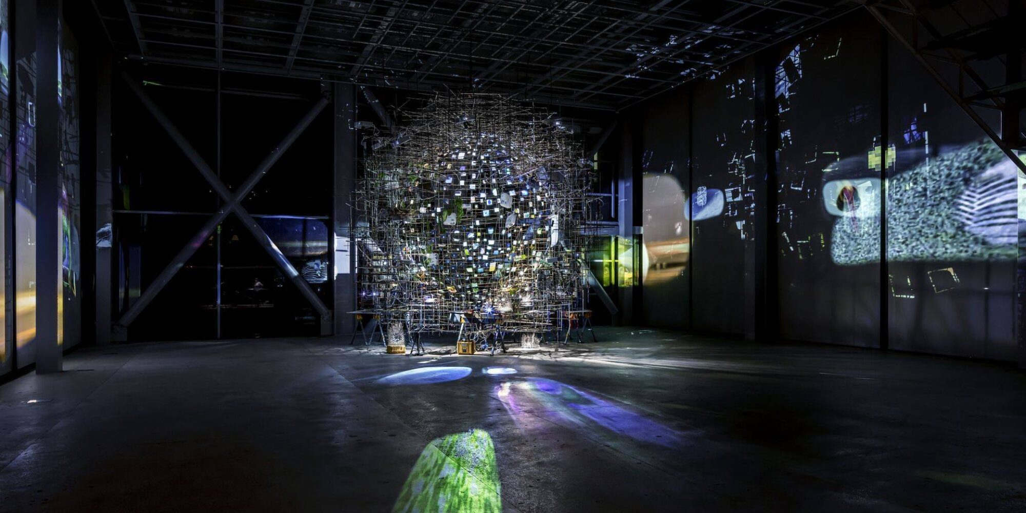 In a black gallery stands an orb-like structure of many small lights surrounded by scaffolding. Large projections of blue and white light project onto the floor, and scenes play on the left and right wall.