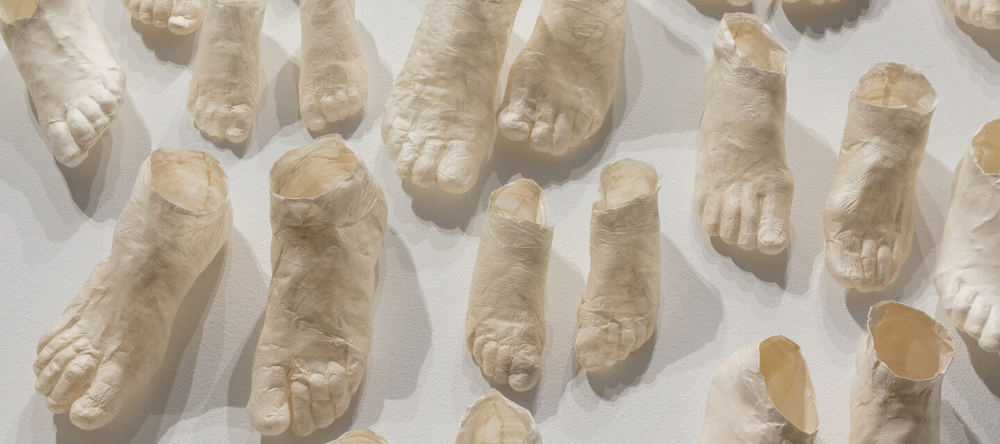 Dozens of pale, hollow, ghostly feet of different sizes made of translucent paper arrayed in pairs on a white background.