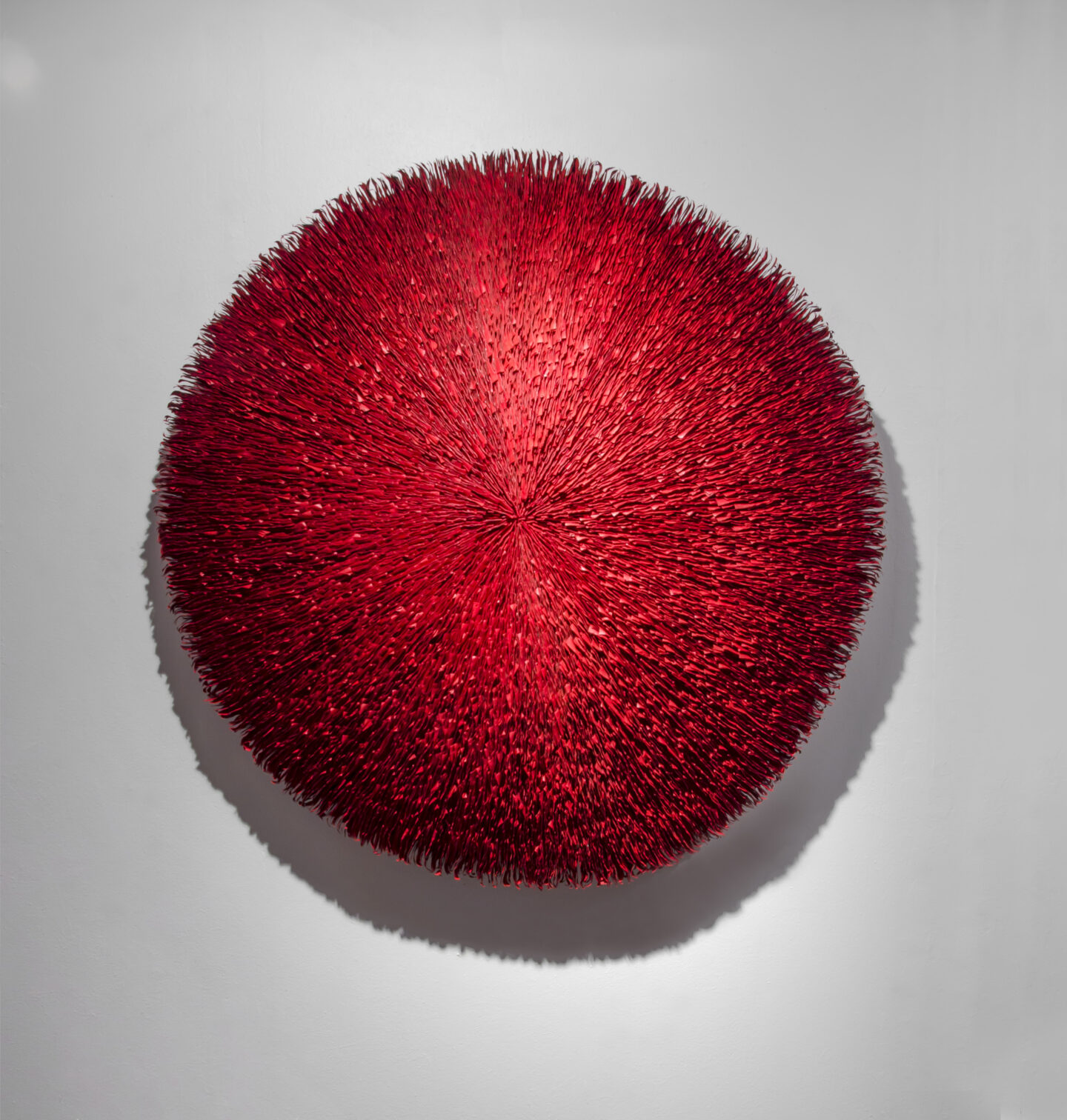 A frontal view of a deep red abstract round wall sculpture made from stacks of paper. Surface is highly textured with lines radiating outwards from the center.