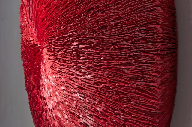 An oblique view of a deep red abstract round wall sculpture made from stacks of paper. Surface is highly textured with lines radiating outwards from the center.