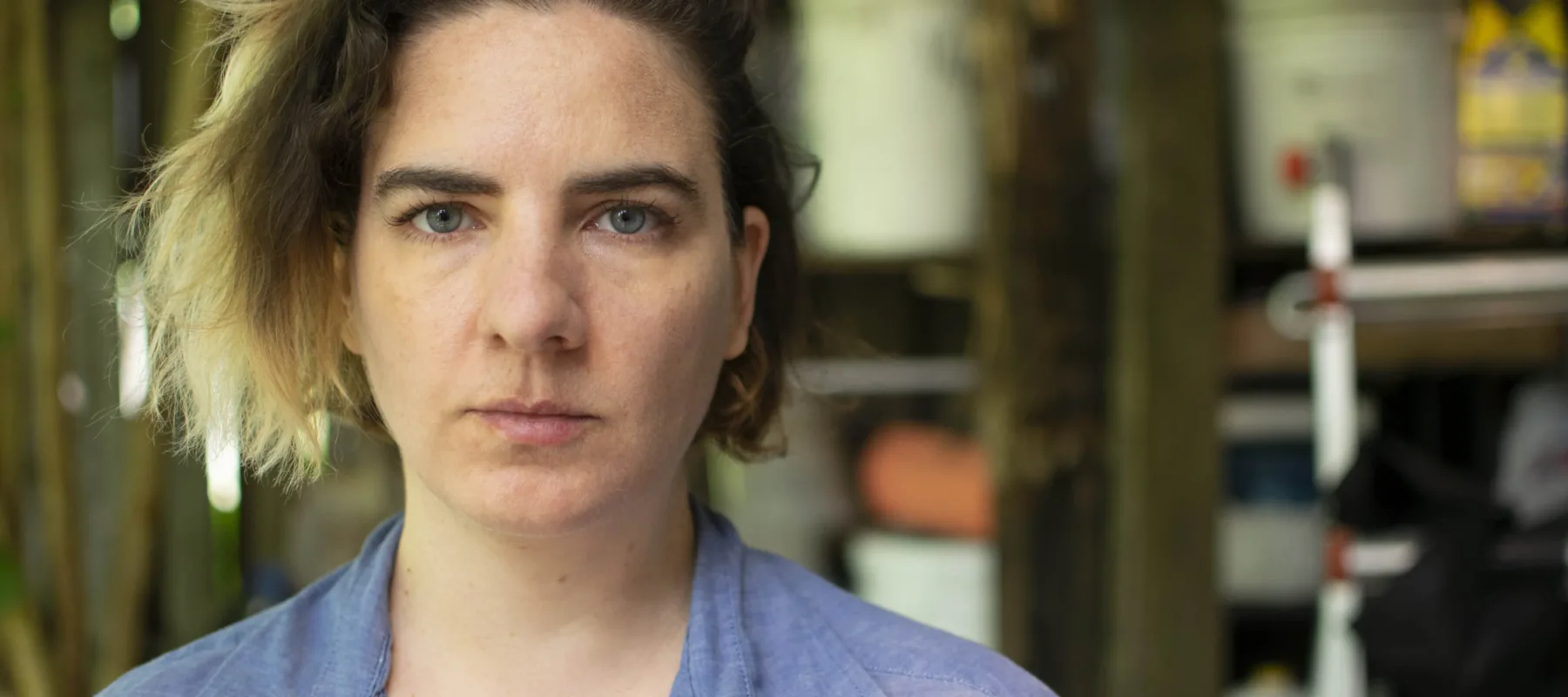 A close up color photograph of a light skinned woman who stares unsmiling at the camera. She has chin-length blonde ombre hair, dark eyebrows, and blue-grey eyes. The background of the photo is blurred.