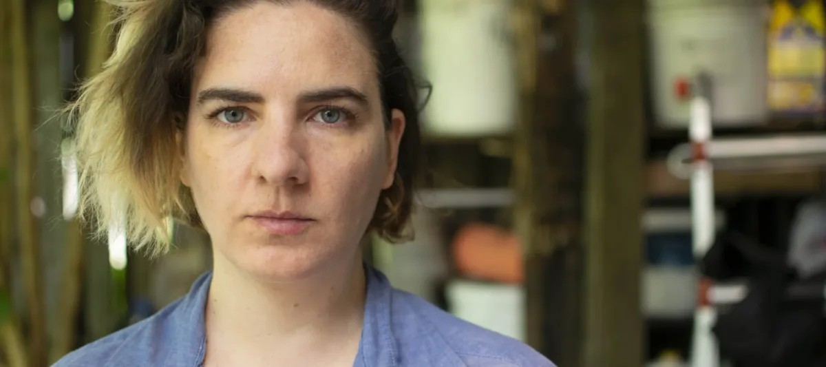 A close up color photograph of a light skinned woman who stares unsmiling at the camera. She has chin-length blonde ombre hair, dark eyebrows, and blue-grey eyes. The background of the photo is blurred.