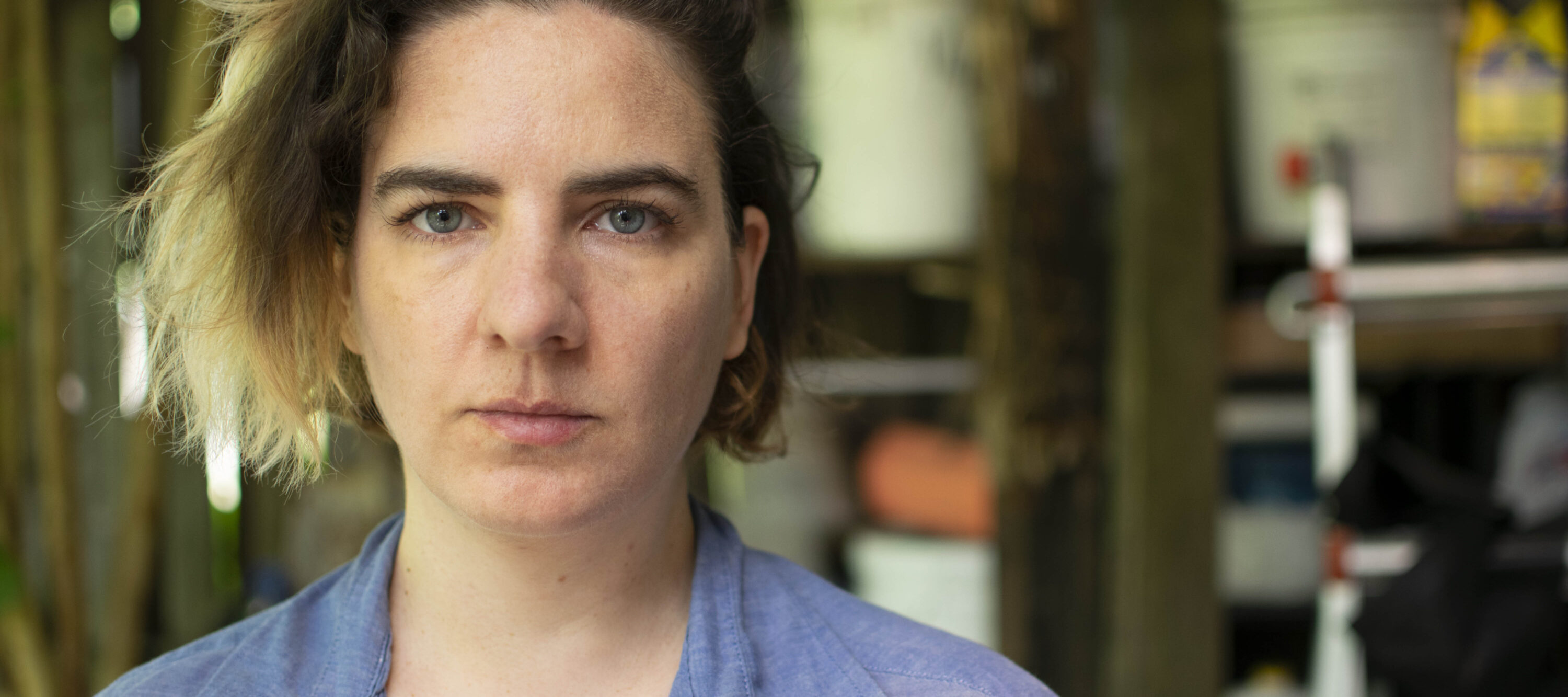 A close up color photograph of a light skinned woman who stares unsmiling at the camera. She has chin-length blonde ombre hair, dark eyebrows, and blue-grey eyes. The background of the photo is blurred.