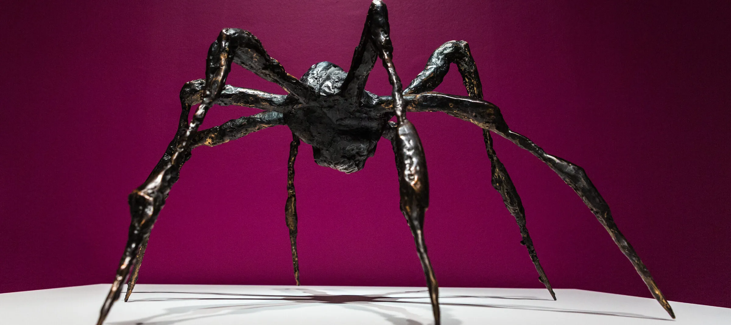 Bronze sculpture of a spider on a white platform against a magenta background.