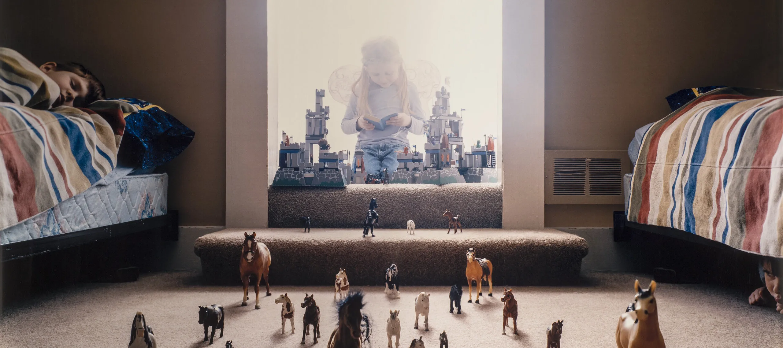 Three light-skinned children: one with long, blonde hair wears fairy wings and kneels, reading a book, behind a toy castle in a doorway full of light; one asleep on a bed on the left; and one under a bed on the right. Between the beds, dozens of toy horses stampede at the viewer.