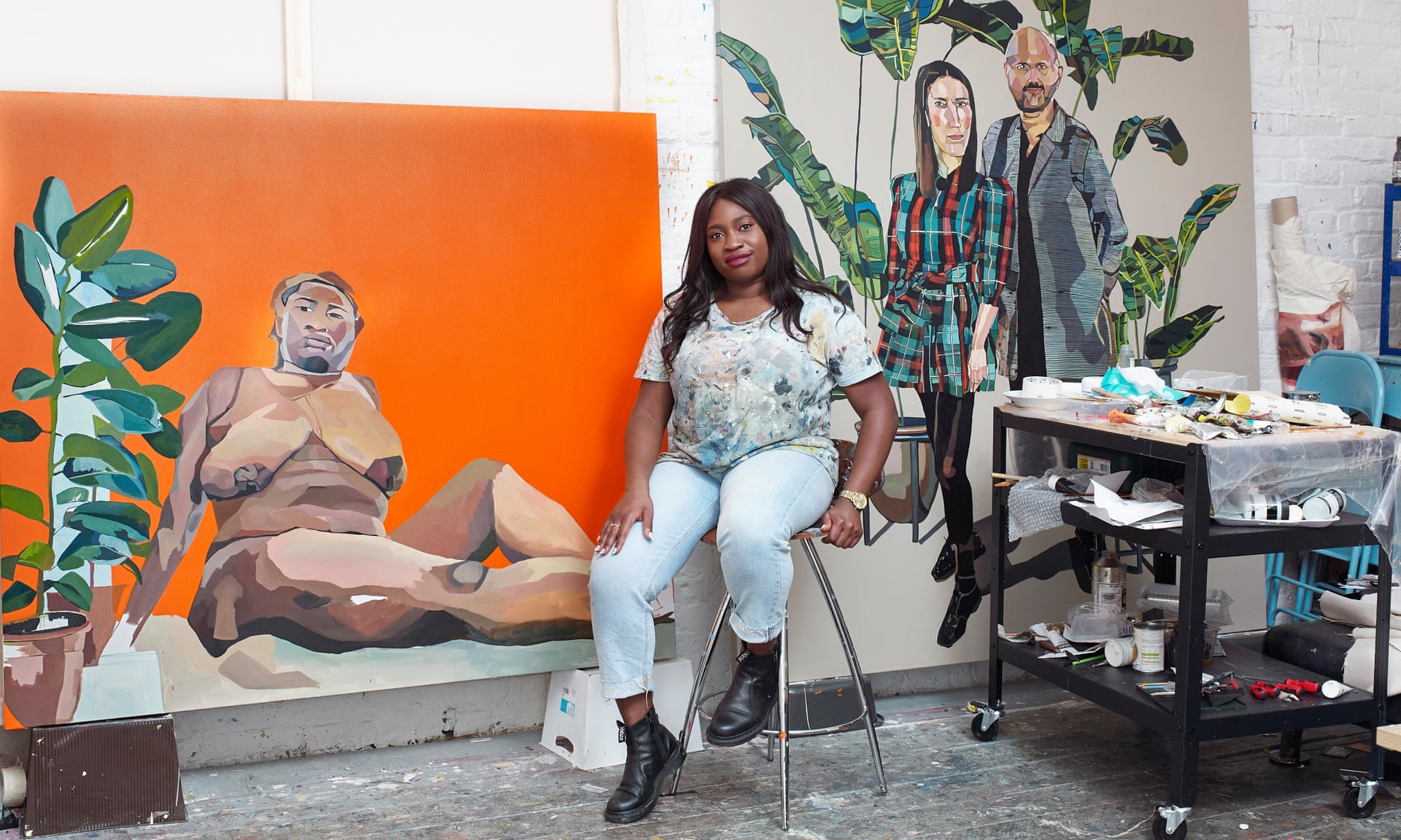 A dark-skinned woman sits on a stoll in an art studio in front of two large portrait paintings. Next to her is a cart filled with painting supplies.