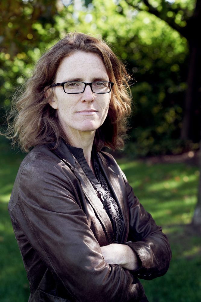 A color photograph of a light-skinned woman with shoulder length brown hair staring directly at the camera unsmiling. She wears glasses and a brown leather jacket and crosses her arms below her breasts. The background is a nature setting, slightly out of focus.