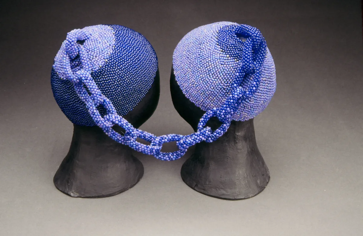 Two head caps made of small, blue glass beads rest on two black mannequin heads. The two caps are connected at the tops by a beaded chain. The left cap is made of darker blue beads and the right cap is made of lighter blue beads. The chain combines both shades.