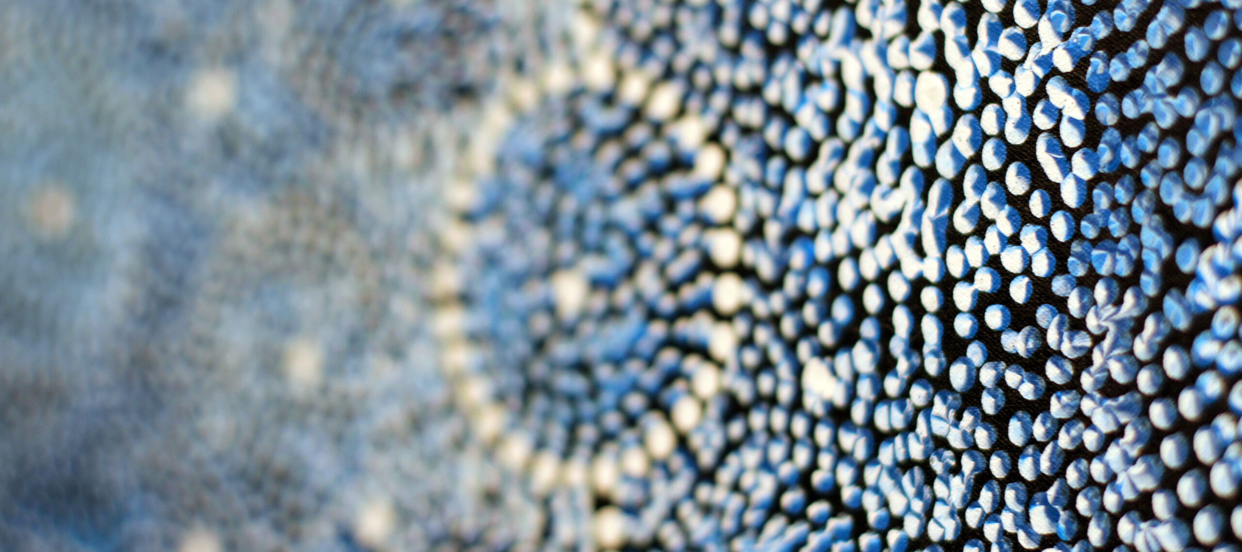 A very close-up detail shot of a painting featuring tiny dots arranged to form vary shades of blue circles outlined in white. Layered blue dots, appearing almost three-dimensional against a black background, are in focus in the foreground and the blue background is blurred.