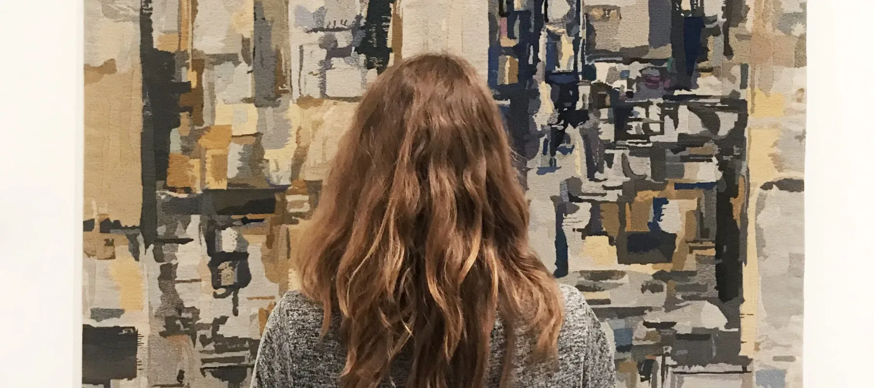 A light-skinned adult woman in a gray dress with medium-length, wavy, brown hair is seen from behind, standing and facing a large, abstract painting. The painting depicts blurry, layered rectangles and squares in shades of gray, taupe, and black, layered and appearing to recede.