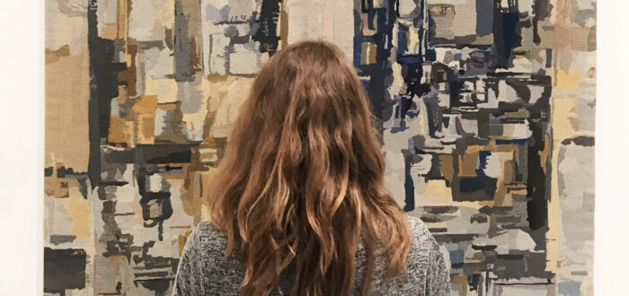 A light-skinned adult woman in a gray dress with medium-length, wavy, brown hair is seen from behind, standing and facing a large, abstract painting. The painting depicts blurry, layered rectangles and squares in shades of gray, taupe, and black, layered and appearing to recede.