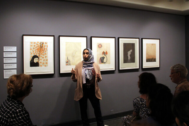 A woman with medium skin tone wearing a multicolored head scarf, beige cardigan, maroon top, and black pants stands in a gallery speaking in front of group of people sitting and listening. The speaker is the artist gesturing while speaking in front of five intricate, detailed prints.