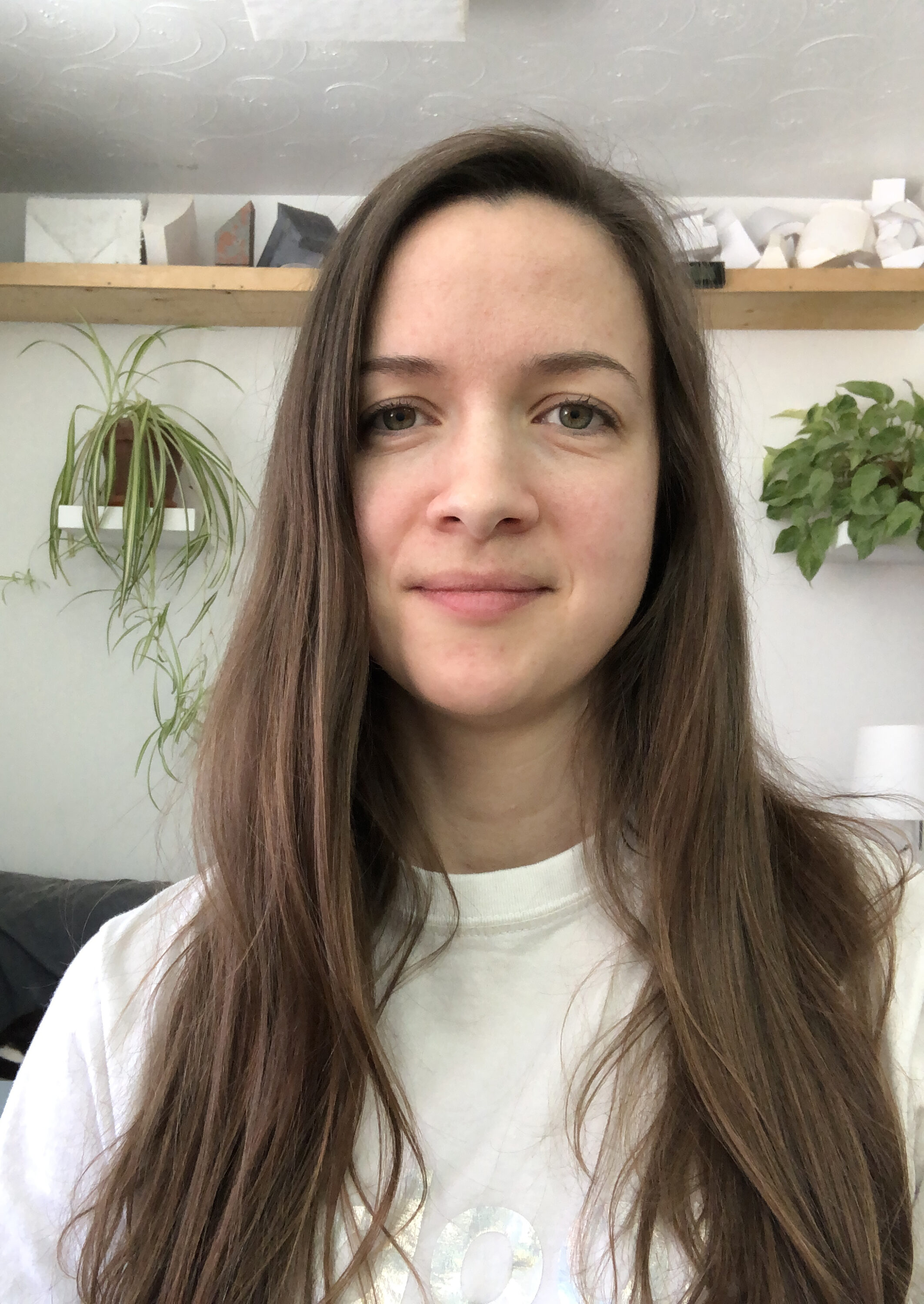 A light-skinned woman with long straight brown hair smiles at the camera. In the background two lush plants are on shelves. The rest of the room is white.