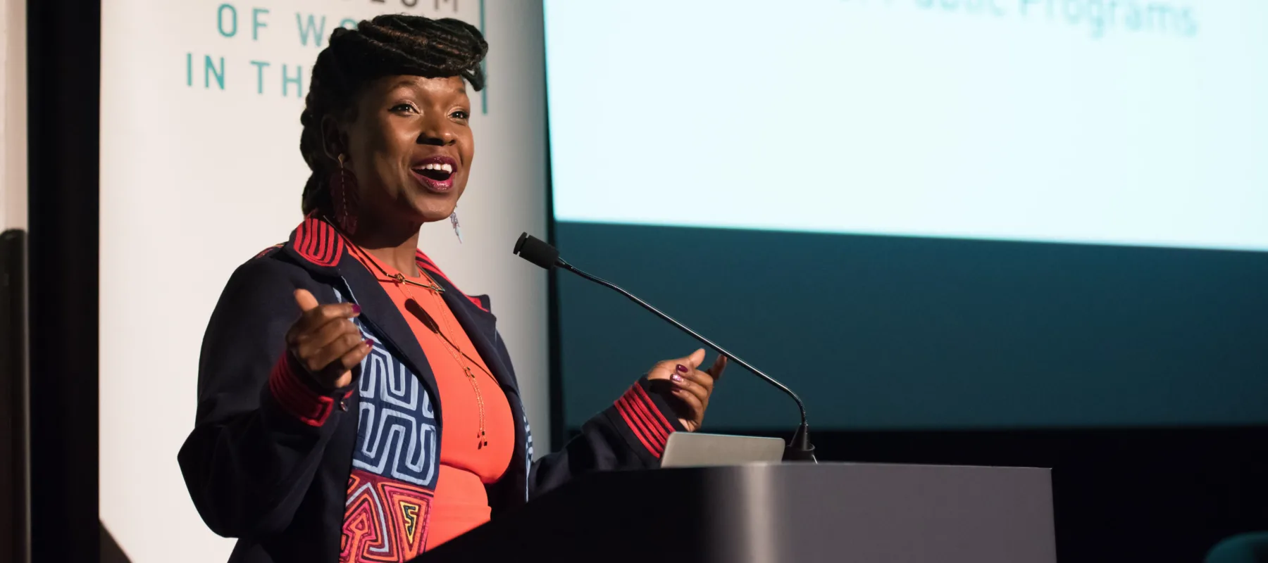 Melani N. Douglass speaks at a podium at the National Museum of Women in the Arts during a Fresh Talk event. She is a dark-skinned adult woman wearing a colorful top and smiling.