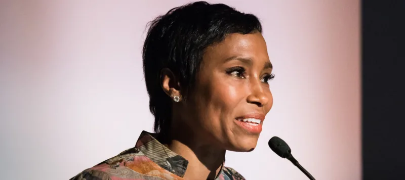 A dark-skinned woman with short cropped hair speaks into a microphone at a podium. Behind her a banner reads "National Museum of Women in the Arts." She wears a colorful abstract print blouse.