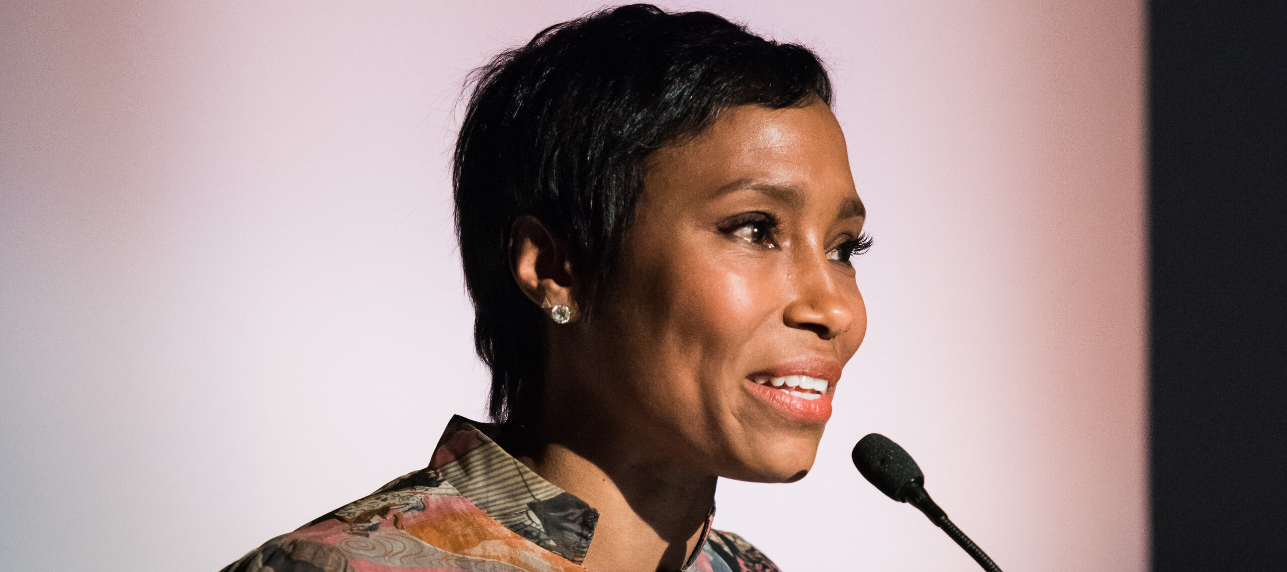 A dark-skinned woman with short cropped hair speaks into a microphone at a podium. Behind her a banner reads 