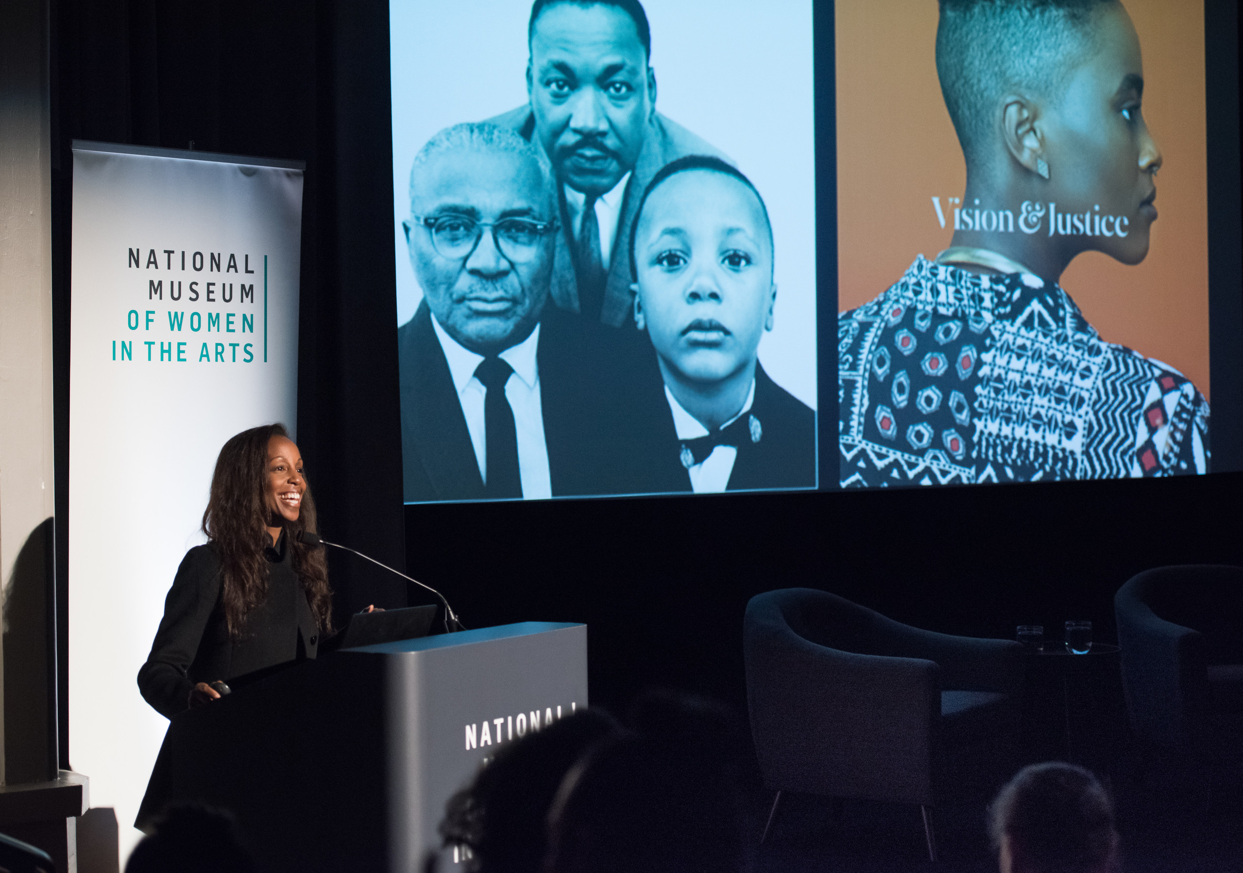 A dark-skinned woman stands at a podium and smiles out at an audience. To her right, on a large presentation screen, are two photographs. One is a black and white portrait of three dark-skinned men including Martin Luther King Jr, the other features a dark-skinned man in profile against an orange background.