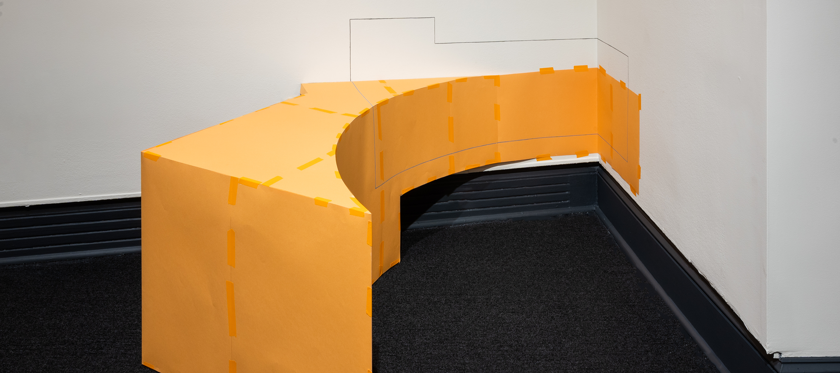 An orange geometric abstract sculpture with varied angles positioned against a corner on the floor. The sculpture has painted lines extending onto the walls.