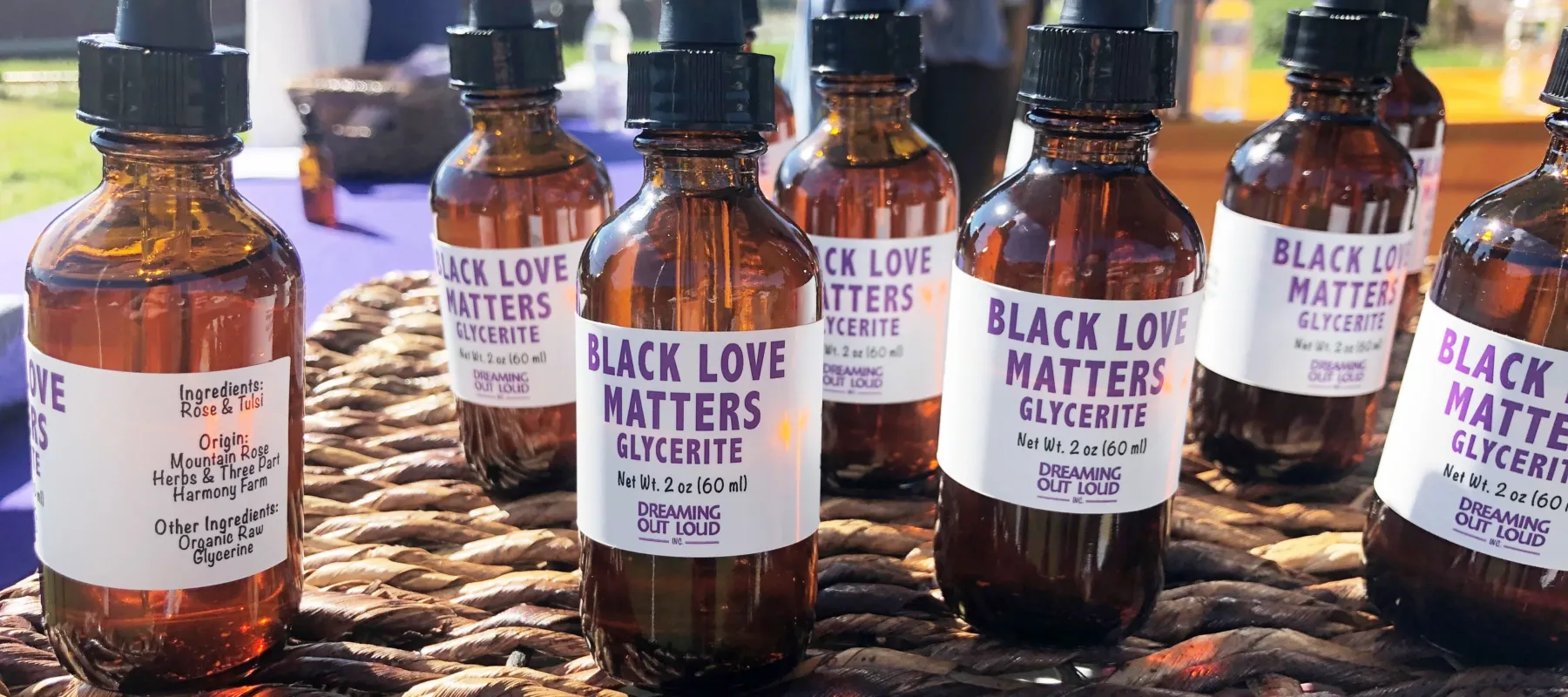 Close-up of several brown bottles with eyedropper lids, each labeled with "Black Love Matters Glycerite." They are perched on a wicker table outside in a field on a sunny day.