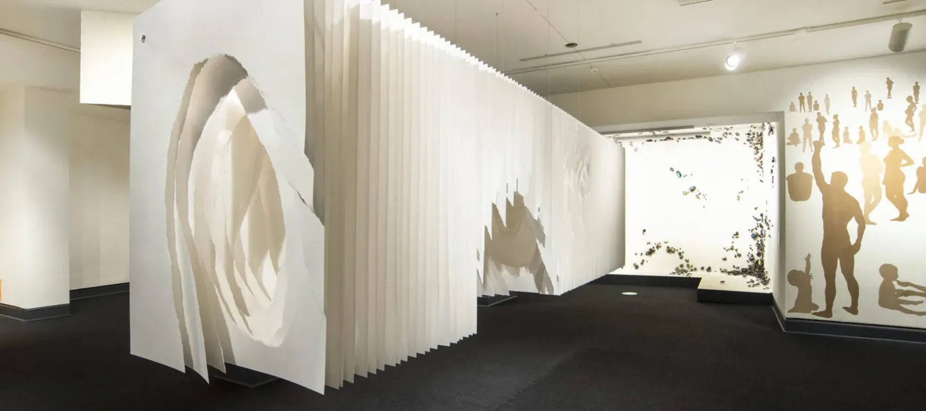 Panels of white paper hung together to look like a long rectangular box suspended from the ceiling a few feet off the ground. Roughly torn holes in the middle of the sheets create a tunnel-like space.