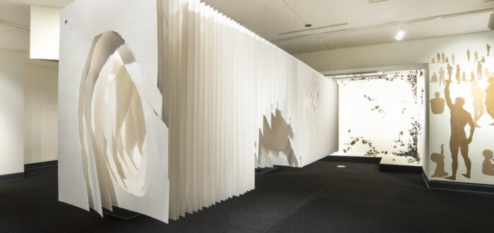 Panels of white paper hung together to look like a long rectangular box suspended from the ceiling a few feet off the ground. Roughly torn holes in the middle of the sheets create a tunnel-like space.