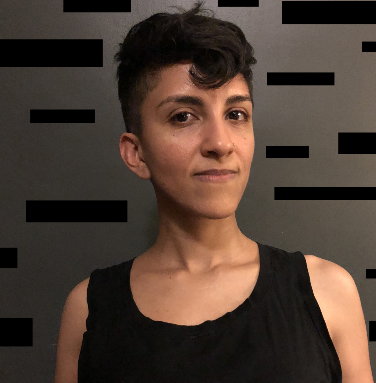 A portrait photograph of a medium-skinned woman with dark cropped hair styled in a pixie cut with one curl swirling over her forehead. She wears a black tank top and looks at the camera with a very slight smile.