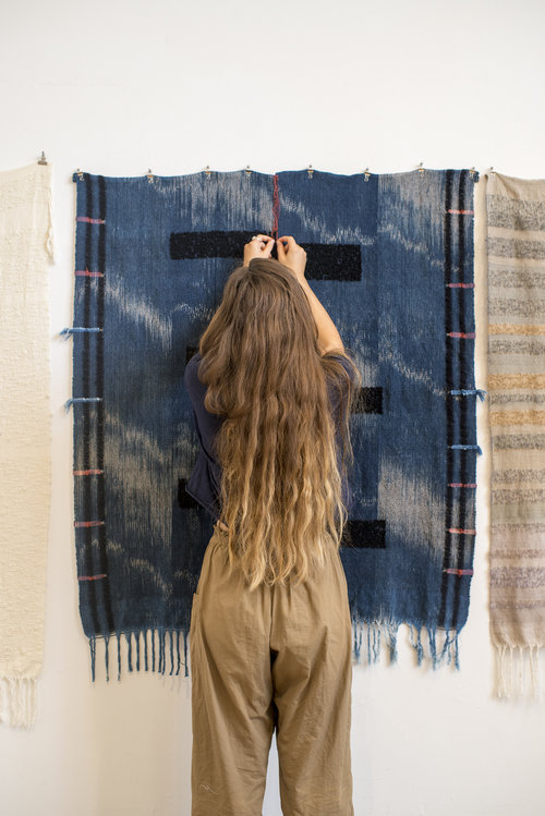 A woman with long, waist-length brown hair stands with her back to the camera and in front of a hanging textile. Her hands touch a red thread, a bit above her height. She wears loose khaki pants and a navy shirt.