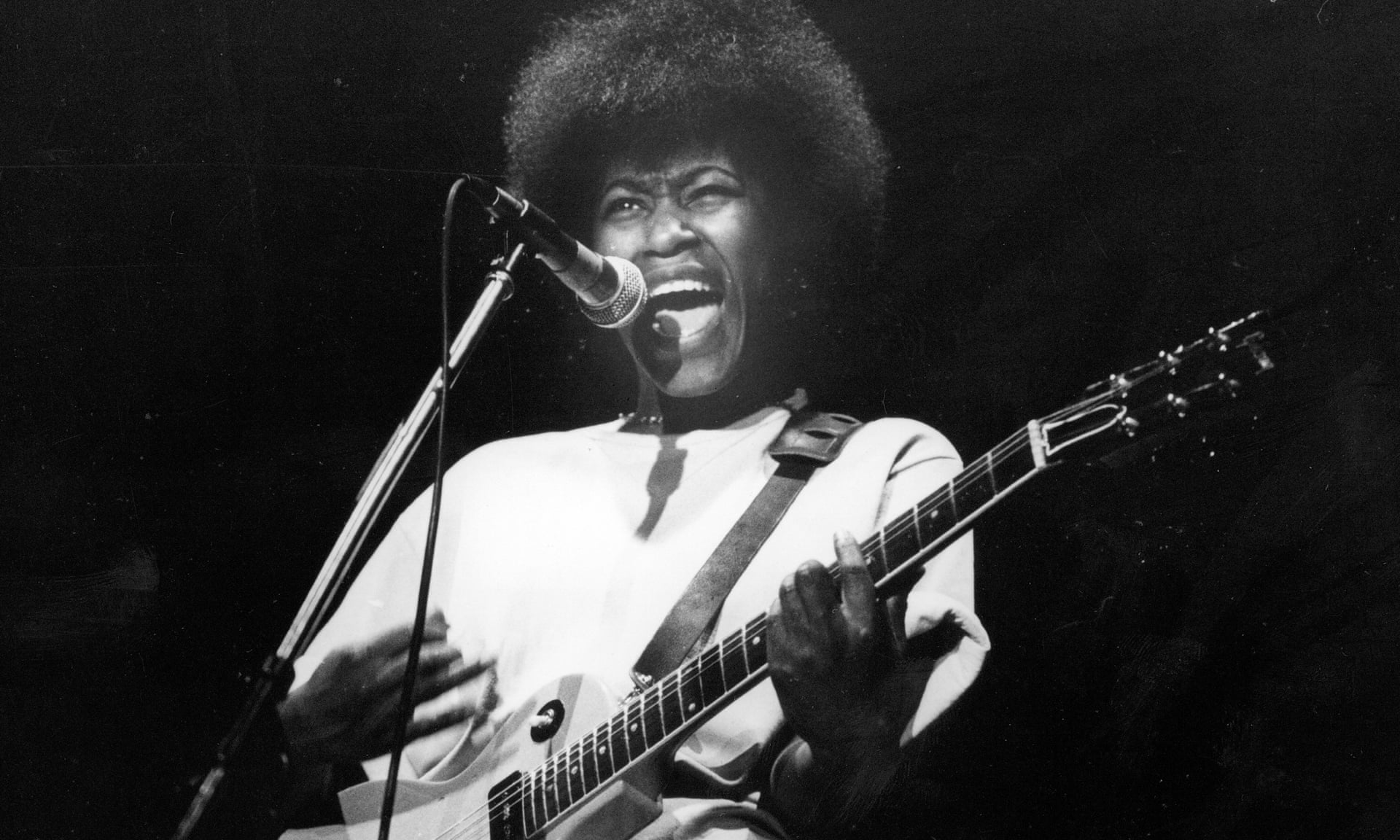 A black-and-white photo of a dark-skinned woman with an afro playing an electric guitar and singing passionately.